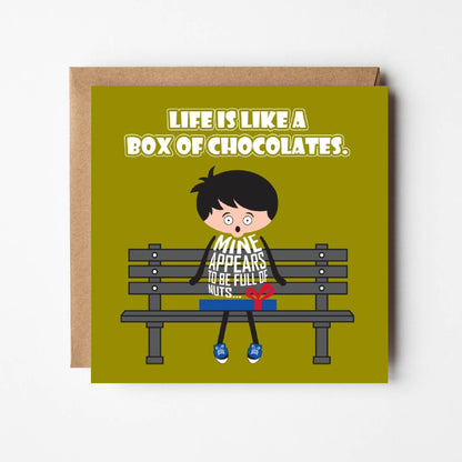 Card titled “Life is like a box of chocolates” in white text on a green background. A cartoon man sits on a park bench with a box of chocolates on his knee just like the the iconic scene from Forrest Gump.  In a reference to both life and his box of chocolates, the man observes that “Mine appears to be full of nuts.” 